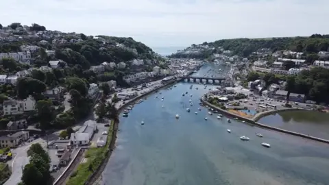 The incident is taking place at the boat yard in Looe. There is a large stream of water running through the middle of the photo. There is the Looe bridge and boats dotted around just infront of it in the boat yard. Either side of the river is greenland, houses and shops.