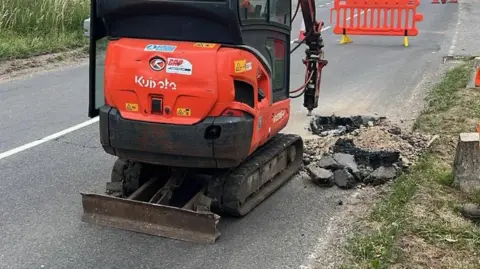 A mobile drill vehicle is parked near a large hole in some road tarmac.