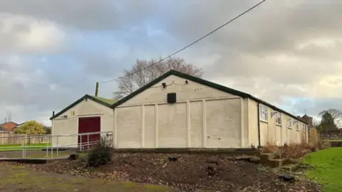 Two beige one-storey buildings with sloped dark rooves surrounded by grass, with a path leading to a red door with metal railings running alongside