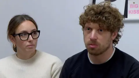 BBC A man with curly brown hair and a black jumper on looking off camera next to a woman with a white jumper on and black glasses. 