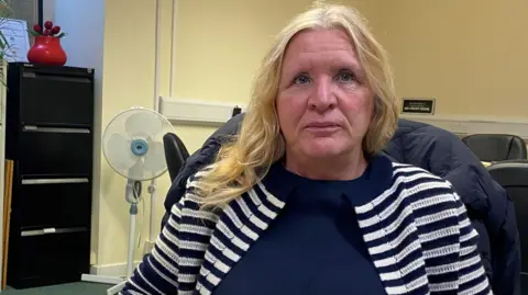 Christine Nicholls is a woman in her sixties wearing a black and white jacket. She has long blonde hair and is staring unsmilingly into the camera 