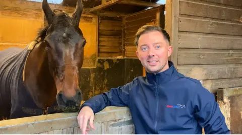 A young man with brown hair and a blue long-sleeve top leaning on the wooden door of a stable, with a brown horse behind the door