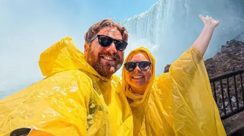 That Travel Couple A selfie of Hannah Bird and Charlie Camper wearing black sunglasses and yellow plastic ponchos. They are smiling and posing in front of Niagara Falls, as water droplets splash down from above.