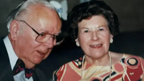 Una Crown and her husband, Ron. He is on the left and almost in profile as if in the middle of talking, wearing glasses with swept back white hair and wearing a dark jacket, white shirt and blue and red bow tie. She is on the right with dark short curling hair, wearing big stud earrings, a pink, red, beige and black patterned dress and a long string of pearls. She is about to smile. 