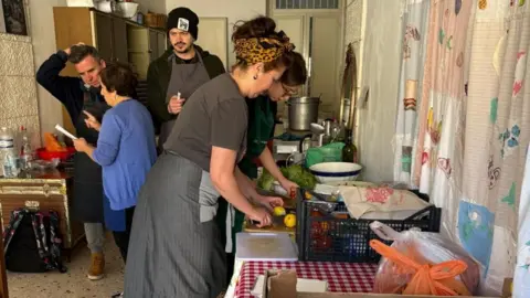 Ingrid is pictured at a workshop cutting some fruit in a kitchen. There is a young chef next to her. Danny McCubbin is talking to a member of staff in the kitchen.