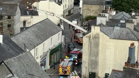Tavistock Fire Station A hydraulic ladder with a team of fire fighters in tis cage is high over the rooftops of houses. There are four fire appliances on the streets below.