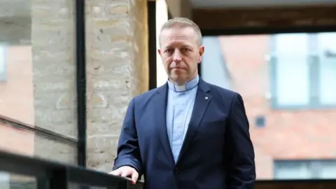 East Belfast Mission Andrew is wearing a minister's collar and a blue shirt, navy suit and he is standing by a window looking at the camera. He has blond short hair and blue eyes. 