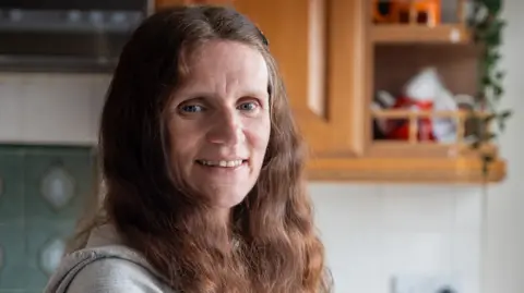 Supplied Wendy Wheat, 53, standing in front of a window, with red, curly hair.