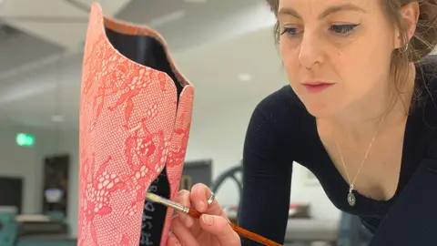 UWE A female designer at UWE applies a paintbrush to the decorated prosthetic. The designer is wearing a fitted black top and a delicate necklace. She looks like she is concentrating.