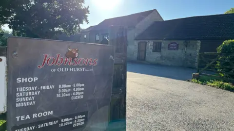 Tom Jackson/BBC Johnsons of Old Hurst sign. Shows opening times. It is a bright sunny morning and there is a brick building in the background . 