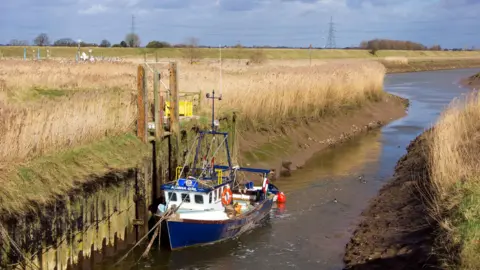 The mouth of the Vernatt’s Drain where it feeds into the River Welland near Spalding, The eastern bank of the Welland can be seen crossing the centre of the image and there is a blue and white boat in the foreground.