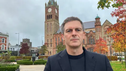 Councillor Shaun Harkin, in a black jacket and sweater, stands in Derry's pece garden. A number of hedges planted in a paved park can be seen in the background as can the city's Guildhall