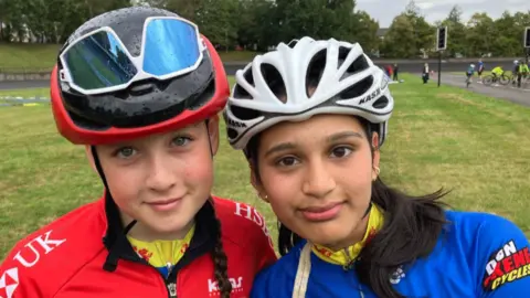 Imogen and Raima in their cycling gear. Imogen is on the left with a red jacket and helmet and shades aloft the helmet. Raima is on the right and has a white helmet and blue jacket on.