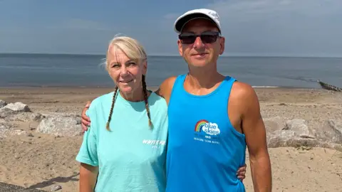 LCC Peter Franklin stands on a beach in the sunshine with his arm round his wife. He is wearing a blue vest top, a white baseball hat and sunglasses and is smiling. Kim is wearing a turquoise tshirt and has blonde hair in plaits