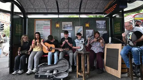 Musicians sat on a bus stop, which has been decorated to appear as a bar. The sign reads "The Metro Sessions". From left to right, a blonde haired girl plays a banjo, a dark haired girl plays a whistle, a brown haired boy plays a guitar, a brown haired boy plays a whistle, a brown ahired boy plays a banjo, a brown haired woman plays a viola and a grey haired man plays a Bodhrán.