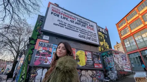 The Canteen Lauren, who has long dark hair and wears a large brown fur jacket, smiles over her shoulder. She is in front of a heavily graffitied wall, with a large billboard for the Canteen behind her. The billboard says: "Bristol, a message from the Canteen: Independent venues are for life, not just for xmas. 24.9% of grassroots music venues are under the threat of permanent closure. Support independent venues before they close for good." 