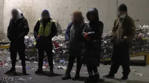 Hungarian police A screengrab from a police video shows some of the men and women arrested in the operation to break up a human trafficking ring. They are standing outside by a rubbish heap, wearing coats, hats and gloves. Their faces are blurred.