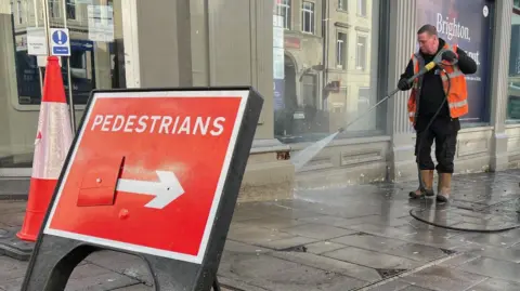 The image shows a sign that says 'pedestrians', and a man can be sen in the back jet-washing the outside of a building.