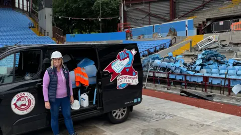 Julie Taylor A woman is pictured standing next to a black van with a side door open. The van features the Scunthorpe United club badge. The woman is wearing a white protective hat. Behind the van is a pile of blue seats ready to be transported from Villa Park to Glanford Park.