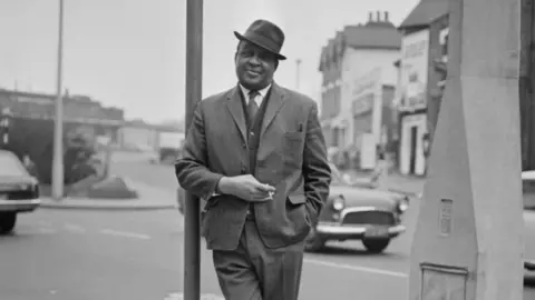 Michael Gains A black and white photo of a man in a suit and hat leaning against a lamppost