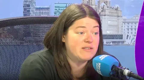 A wide shot of police and crime commissioner Emily Spurrell. she has long brown hair and is sitting in front of a blue microphone labelled 'BBC Radio Merseyside'. Behind her is a screen with a picture of Liverpool's waterfront on.
