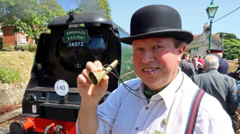 A smiling Peter Sills posing for the camera and holding up the brass horn, which is the size of a large cigar, between his thumb and fingers. He is wearing a white shirt and cravat and a black bowler hat. Behind him is a black and green steam locomotive with Swanage Railway 34072 written on the front.