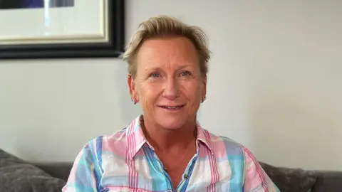 Tara is seated on a grey sofa poses for a portrait in a living-room setting. She is wearing a pastel check-pattern shirt, with framed artwork hanging on the wall behind and cushions on either side.