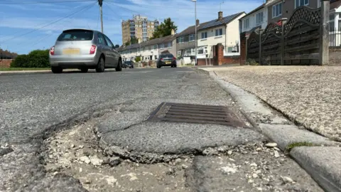 A pothole and crumbling pavement on a street of 1960s council houses, with one of Penhill's tower blocks in the background.