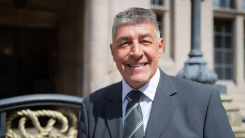 Staffordshire County Council Councillor Chris Large, a man wearing a grey suit and white shirt. He is standing outside Stafford County Buildings and is smiling for the camera.