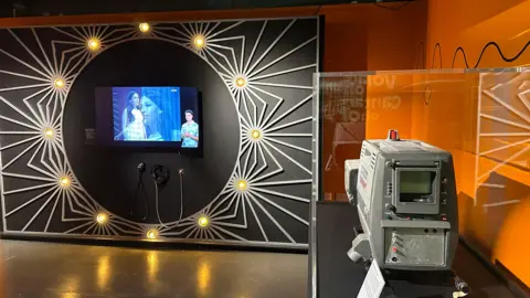 A screen showing Eurovision highlights with BSL interpretation on a wall with lights in a circle around the screen. To the right of the picture there is an old BBC TV camera head in a glass case. The walls are a bright orange.