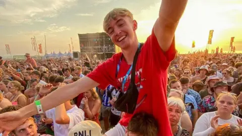 Jane Johnson Close-up of a large outdoor festival crowd at sunset. In the foreground, one person in a red T-shirt sits on another person’s shoulders with arms raised, wearing a small black crossbody bag. Around them, a large crowd stands closely packed together. A stage structure is visible in the background, along with flags and a partly cloudy sky illuminated by warm evening light.