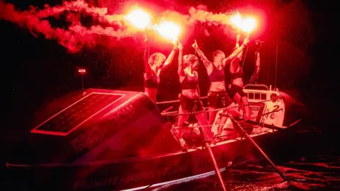 Imy O'Brien, Becca Glover, Sheri Lucas and Emma Gibb stand on their 8.5 meter row boat Rosie. They have are celebrating completing their 3000 mile journey. It is night time and they are all standing with their arms aloft holding red flares which are lit and providing the only light in the photo. Two oars are visible in the water in front of them. 