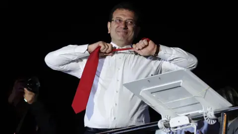 Epa Ekrem Imamoglu, vestindo uma camisa branca e sorrindo, remove uma gravata vermelha