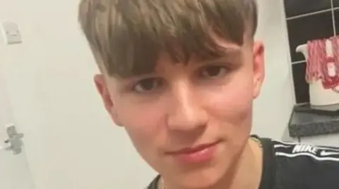 A selfie of a teenage boy who has a half-smile, with fair hair in a fringe cut straight across his forehead. He is wearing a black Nike top and appears to be in a kitchen, with a kettle, black tiles and white wall in the background.