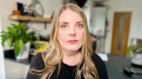 Nikki Fox/BBC Chloe Longster's mother Louise Longster looks at the camera in her kitchen. She wears a black, long-sleeved top and has long, dark blonde hair and blue eyes.