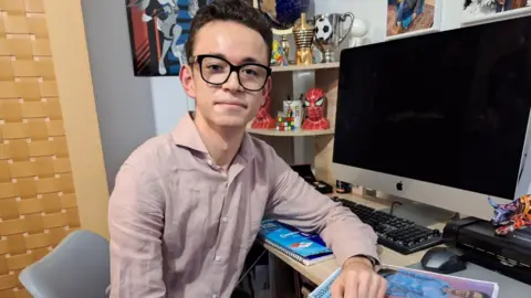 BBC Kenan sitting at a desk. His arm is resting on his own manifesto and he is looking into the camera. There are trophies and a Spiderman ornament on a shelf behind him.