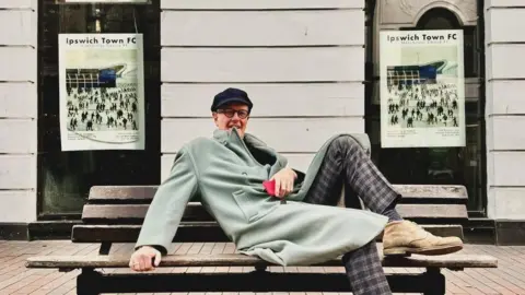 Call Me Ted Simon Farr, lying down on a bench, with his foot up, wearing checked trousers and a green coat, between two Ipswich Town posters