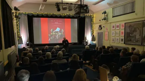 Lucem House Community Cinema An internal view of the Lucem House Community Cinema with several rows of people sat in blue cinema seats looking towards the screen which is showing a graphic for the film Conclave