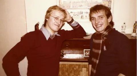 Mayall Family An old photograph of wo young men standing in a room. On on the left, a young Ade Edmondson has light brown or blonde hair and glasses and is resting his elbow on a sideboard. He is wearing a shirt underneath a red v-neck sweater. On the right, a young Rik Mayall has darker hair and is wearing a dark sweater with a tartan scarf. They are smiling at the camera.