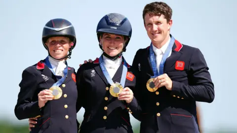 PA Media Rosalind Canter, Laura Collett and Tom McEwen. Rosalind and Laura are wearing horse riding helmets, whilst Tom is not. The trio are all dressed in equestrian gear and wearing medals around their necks. They have their arms around each other and are smiling 