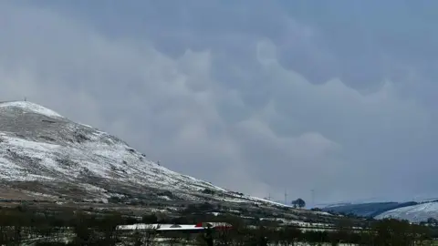 Rachel Cassidy A photograph of a winter landscape taken at the Sperrins on 20 November 2024. Snow and frost can be seen on the ground. A large hill also topped with snow is to the left of frame. A building can be seen among leafless trees below the hill. The sky is grey and cloudy and covers much of the frame. Three wind turbines can be seen faintly in the background. 
