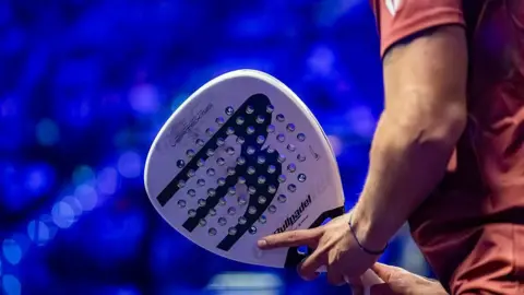 A close up of a man wearing a red t-shirt holding a padel. It is similar to a tennis racquet but is thicker and white, with dozens of tiny holes on its surface. It is white with black writing