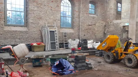 The inside of a chapel which has stone walls and large windows. The inside is a building site with a cement mixer, digger, bricks and other building supplies.