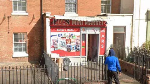 The front of the Mars Mini Market convenience store. It's a small shop set back from the pavement behind some metal railings. The main window is covered in product adverts and then is doorway with an open door. There is a person dressed in leisurewear walking towards the entrance. It reads Mars mini market across the shop front and there is a white retracted canopy above it. 