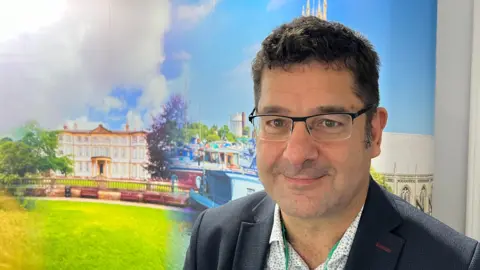 Amanda White/BBC A man wearing a dark grey jacket and black-rimmed glasses. He is standing in front of an image featuring a large ornate building and a church.