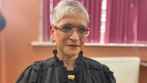A woman with short grey hair and glasses, wearing a black blouse with a gold brooch, stands in front of a coral coloured wall and a window with pink curtains.