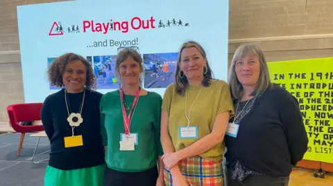 Four women pose for a photo in front of a projector screen with the words Playing Out and some pictures of children playing outside. A large banner with writing on it is just visible to the right.