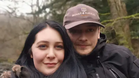 Contributed A young woman with long dark hair takes a selfie in a woods with a young man. She wears a green coat with a fluffy hood. The man wears a purple cap with a black coat. They are both smiling. 