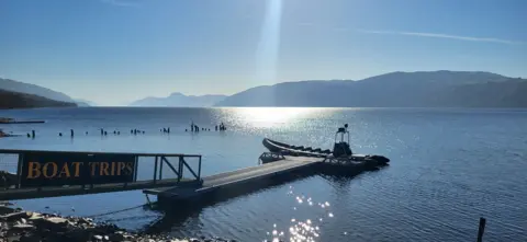 Emma Watson Loch Ness on a sunny day - the sun is shining down and reflecting on the water. A boat sits by the dock, with a sign saying BOAT TRIP on the walkway leading to it
