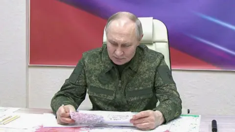 Putin sits at a table reading from a piece of paper with handwriting on. In the background is a painting with red and purple colours. He is wearing combat gear